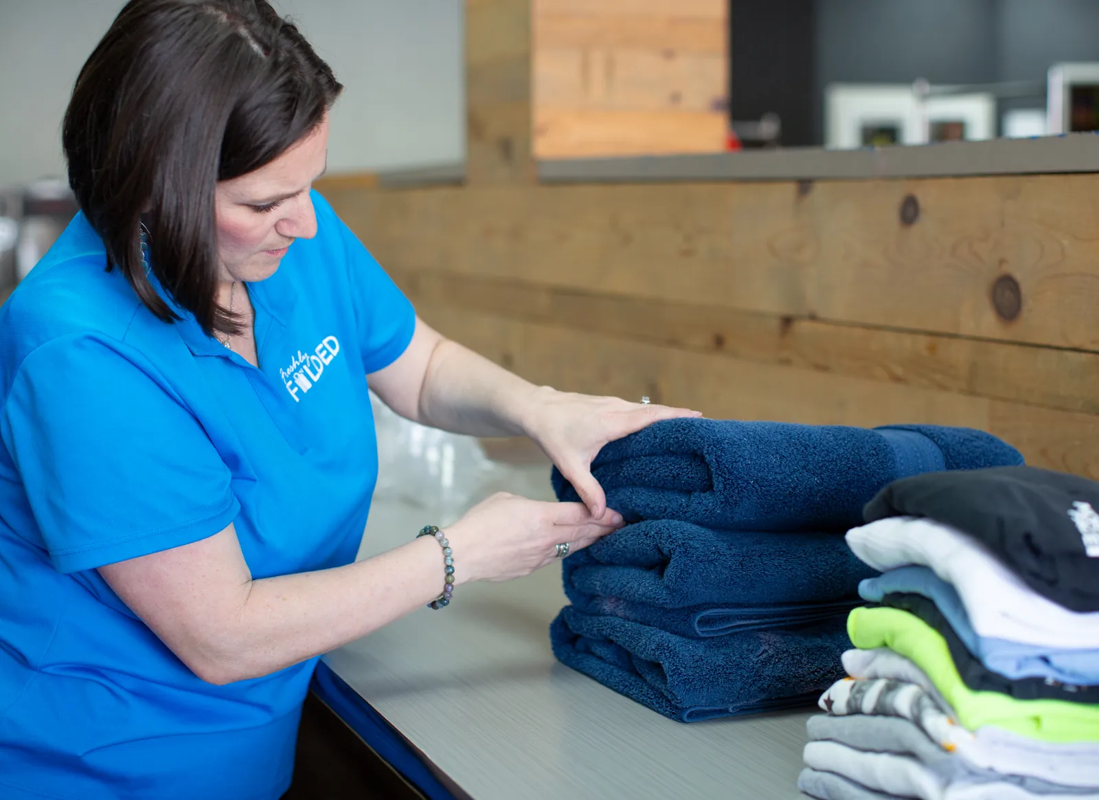 A Freshly Folded team member folds navy bath towels on a work table beside a stack of folded shirts.